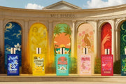 Five perfume bottles displayed in front of a grand classical building facade with colorful illustrated panels behind them. The bottles are labeled Mes Bisous, with scents including Advanced Dream, Tropical Paradise, Petite, Passion, and Green Tea.